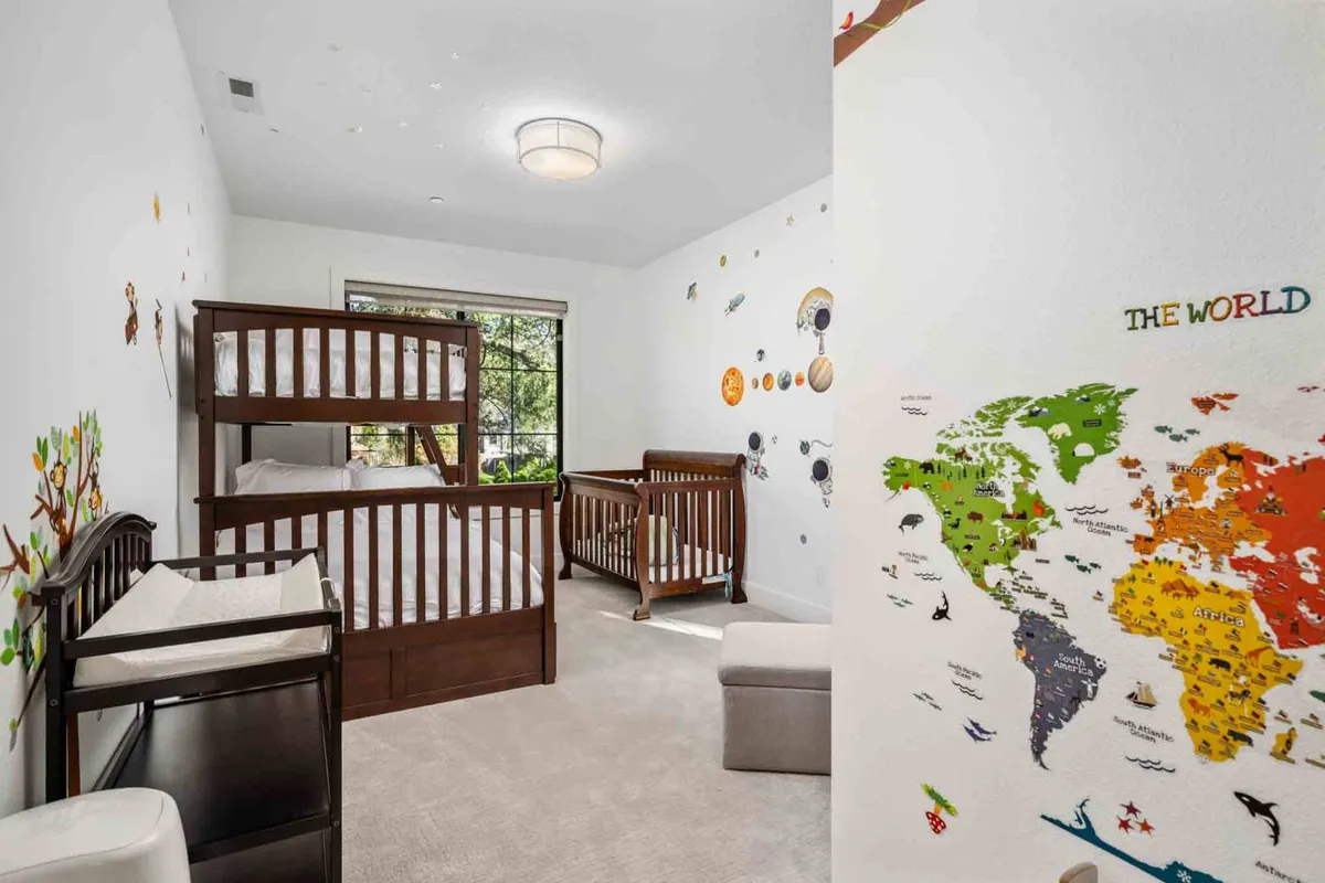 A bright and cheerful bedroom featuring twin-over-full bunk beds, a crib, and playful wall decor with a world map. Natural light pours in through the window, lighting up the space perfect for families or children.