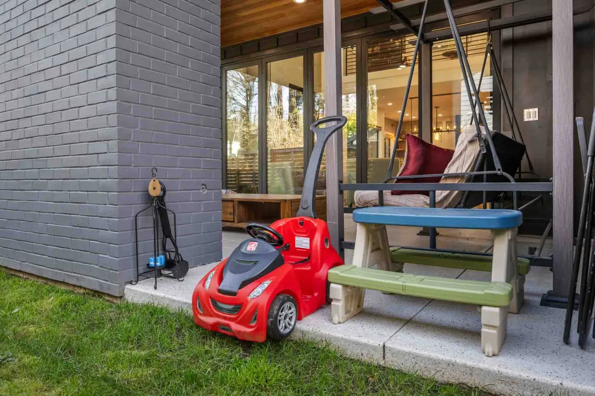 An exterior view of a fully fenced yard features a red toy car, green and blue bench, and a swing seat. The scene is playful and safe, inviting children and pets to roam and explore within the landscaped and enclosed outdoor space.