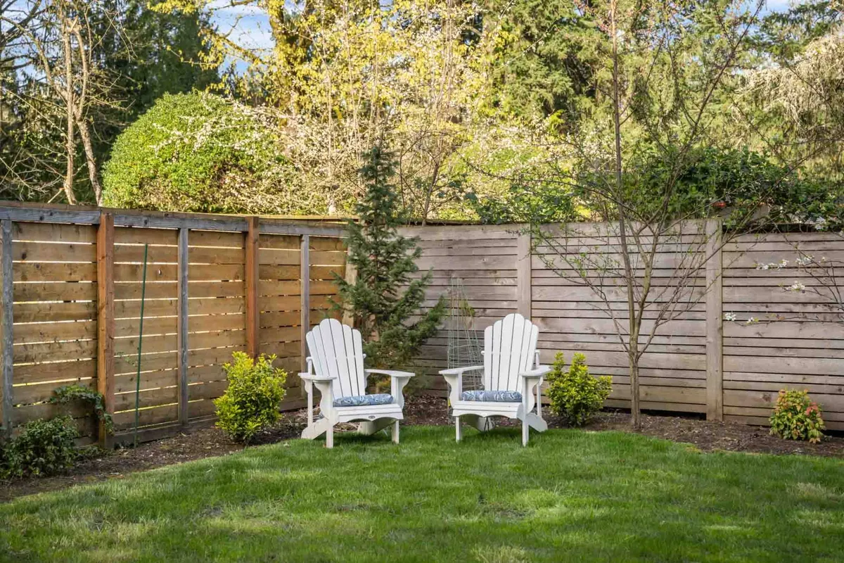 A peaceful outdoor seating area with two white chairs on green grass, bordered by a wooden fence and lined with small trees. The space exudes relaxation, ideal for morning coffee or evening conversations.