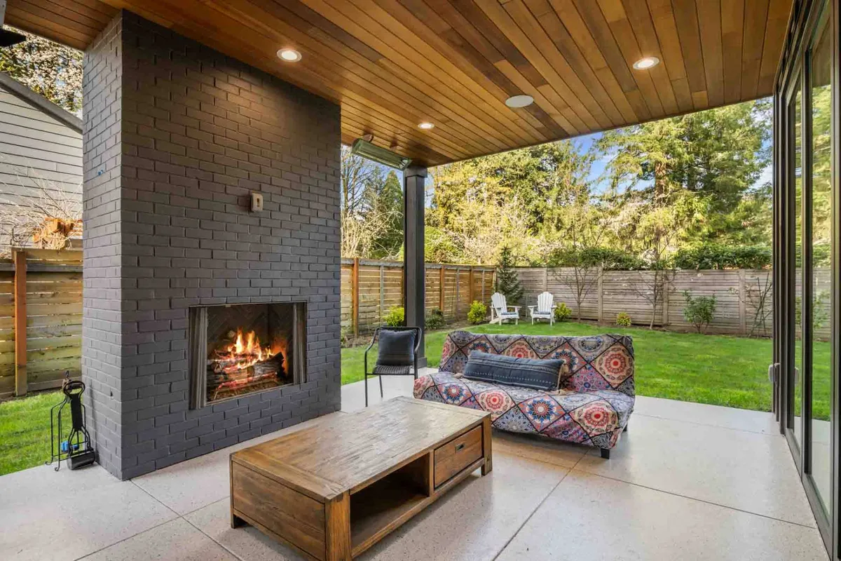 An inviting outdoor seating area featuring a cozy sofa, a modern fireplace, and a coffee table, all set on a patio surrounded by greenery. The space is staged for relaxation, with the fire feature suggesting warmth for evening gatherings or early mornings.