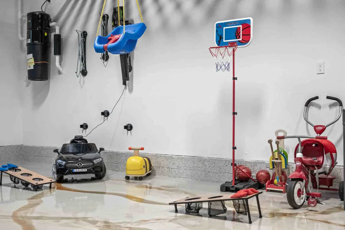 This image displays a playful, energetic interior scene with a toy basketball hoop, kids' toy car, swing set, cornhole boards, and a tricycle. The space is tailored for children's enjoyment with plenty of open area for play and games.