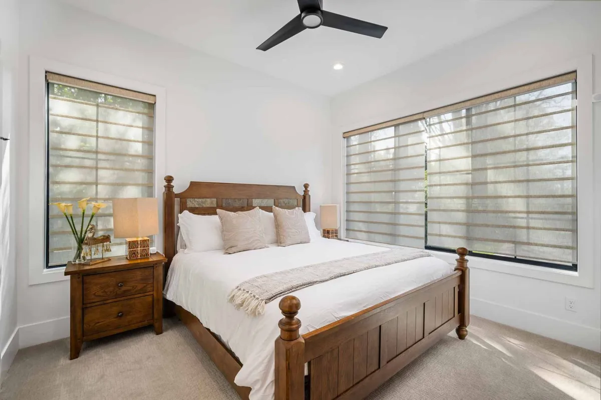 A serene bedroom with a wooden bed frame adorned with decorative pillows and flanked by table lamps. Soft natural light filters through the window treatments, creating a tranquil and cozy ambiance ideal for relaxation or reading.