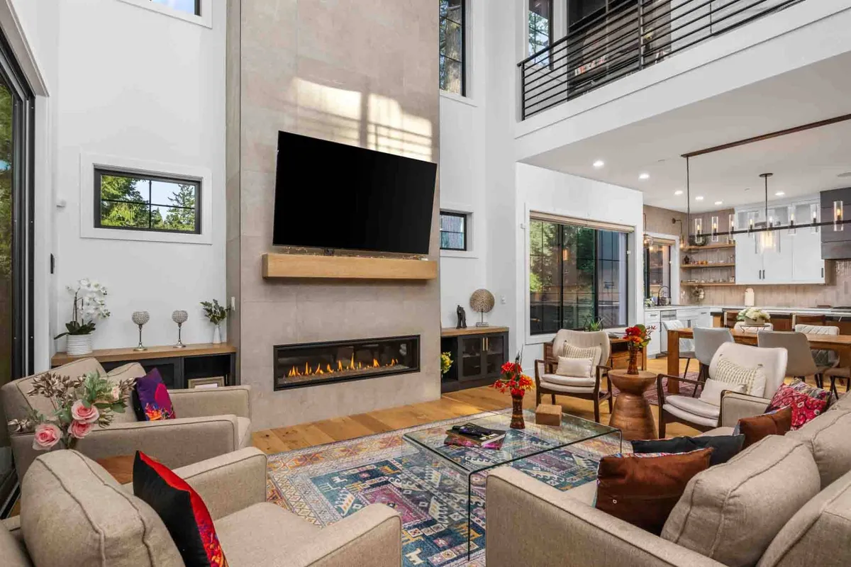 The image showcases an inviting living room featuring a glowing fireplace as the centerpiece, flanked by large windows that let in ample natural light. A colorful rug anchors a plush seating arrangement, creating a warm and stylish setting perfect for gatherings. A sleek TV and upscale décor complete the scene, emphasizing comfort and sophistication.