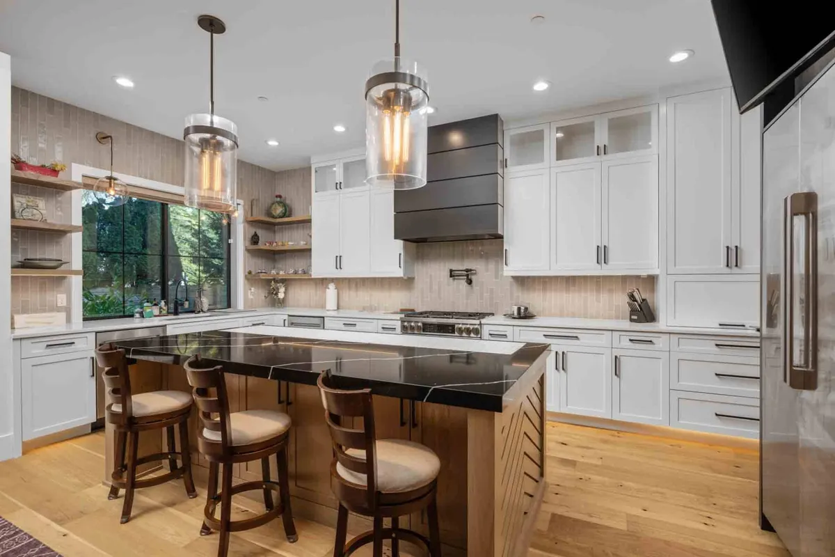 A spacious, modern kitchen featuring a central island with a striking black countertop, white cabinetry, and stylish light fixtures. Large windows bring in lush greenery and natural light, creating a bright and inviting atmosphere perfect for cooking and entertaining.